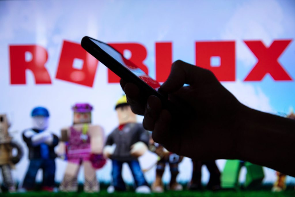 Особа држи паметни телефон док се у позадини види лого игре Роблокс (Roblox) са фигурицама